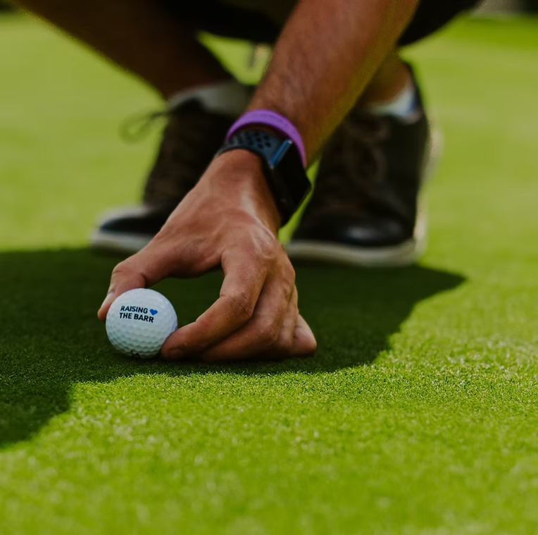 A man places a golf ball on a putting green. The ball is labeled with the words "Raising the Barr."