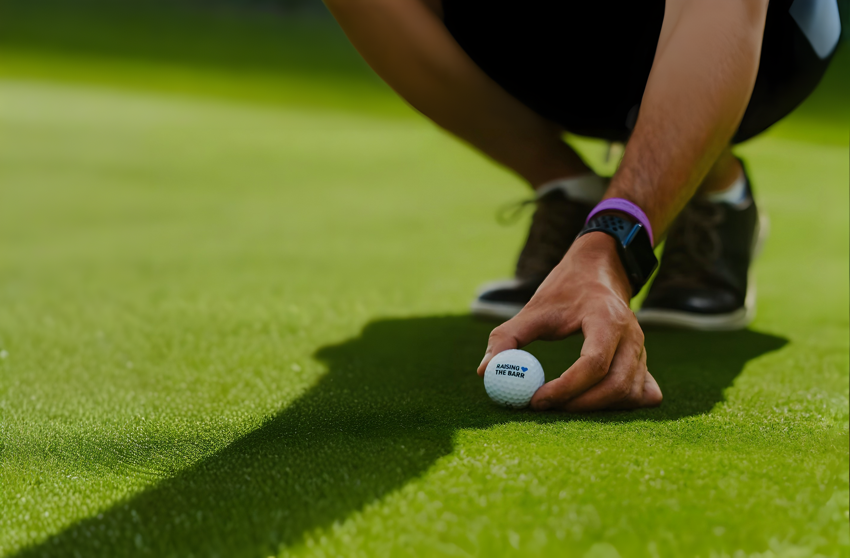 A man places a golf ball on a putting green. The ball is labeled with the words "Raising the Barr."